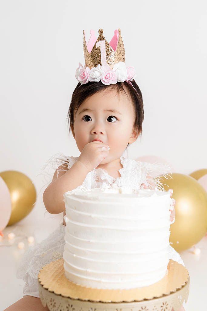 Baby girl in white dress and gold crown smiles while tasting her first birthday cake surrounded by gold balloons.