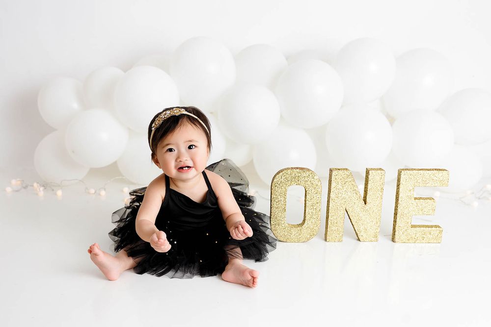 Baby girl in black tutu sits smiling beside gold glitter ONE sign with white balloons for first birthday.