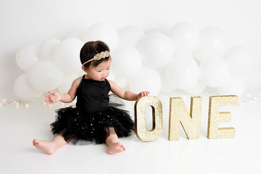 Baby girl in black tutu sits beside gold glitter ONE letters with white balloons for first birthday.