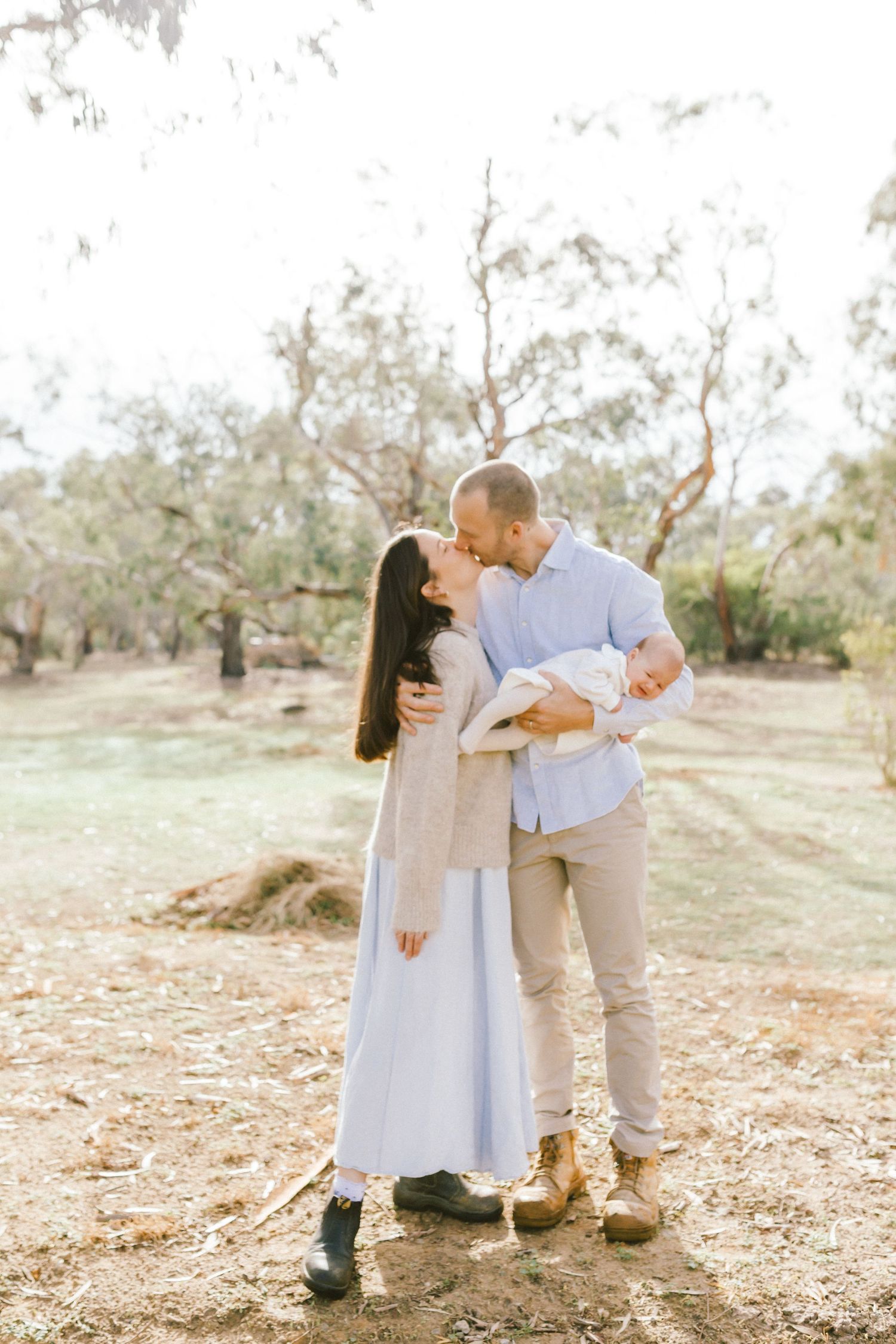 A couple kisses outdoors in a sunlit park while the man holds their newborn baby, wearing light blue and neutral tones.