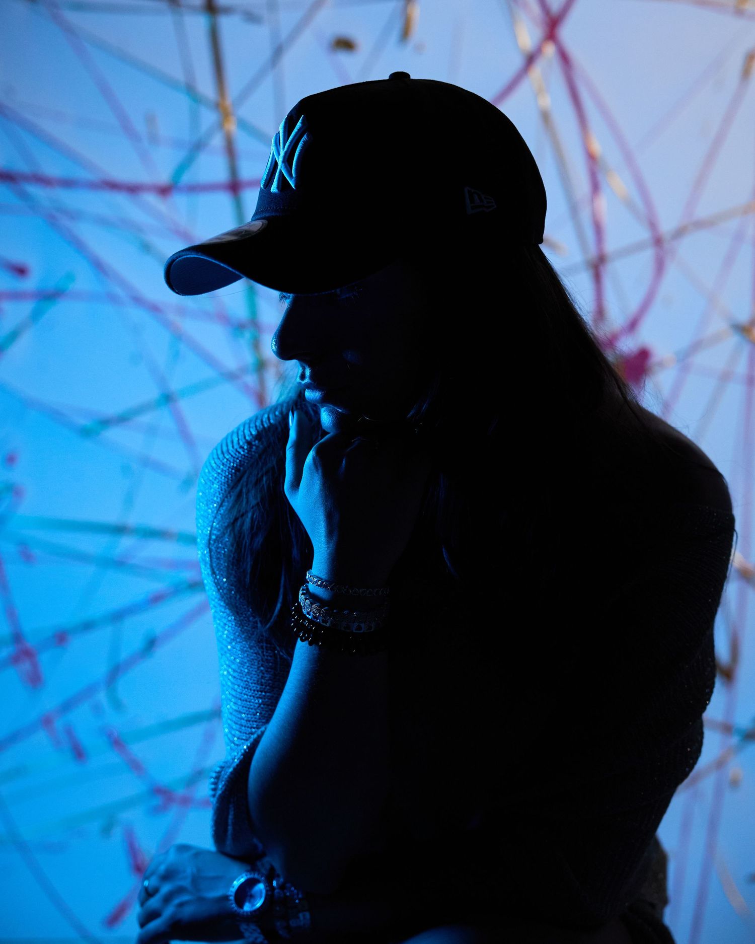 Person in black NY Yankees cap poses thoughtfully in blue-lit studio with colorful abstract background.