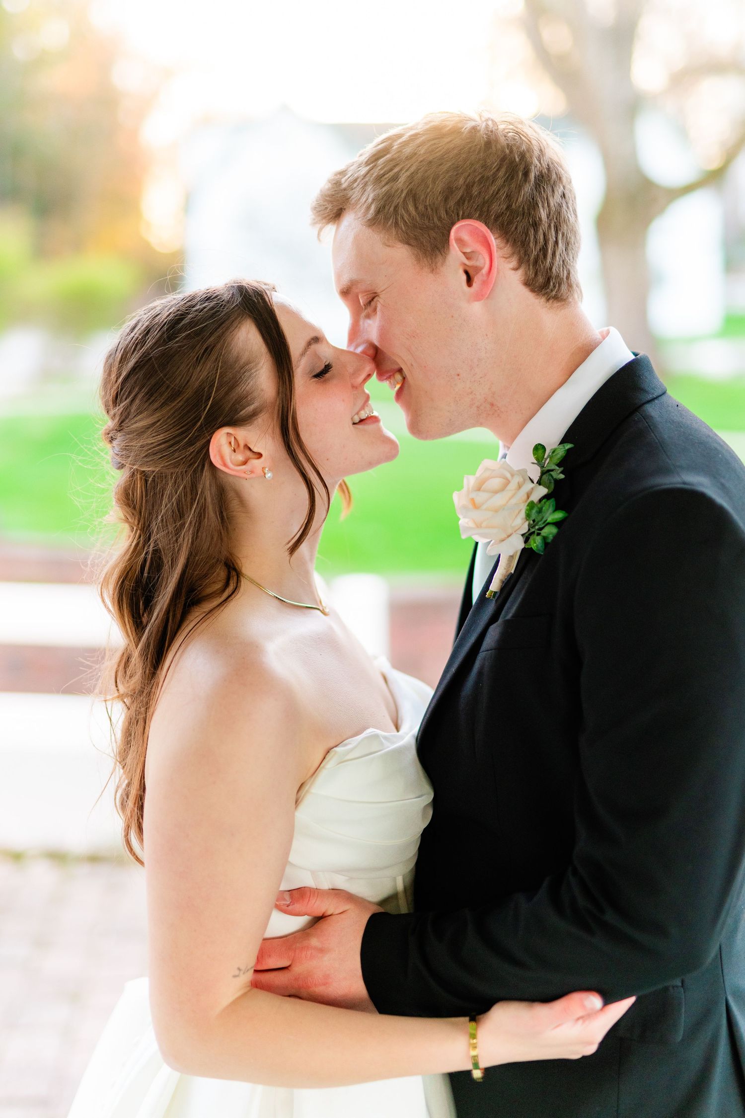A bride and groom share a romantic almost-kiss moment, dressed in wedding attire with a white rose boutonniere.