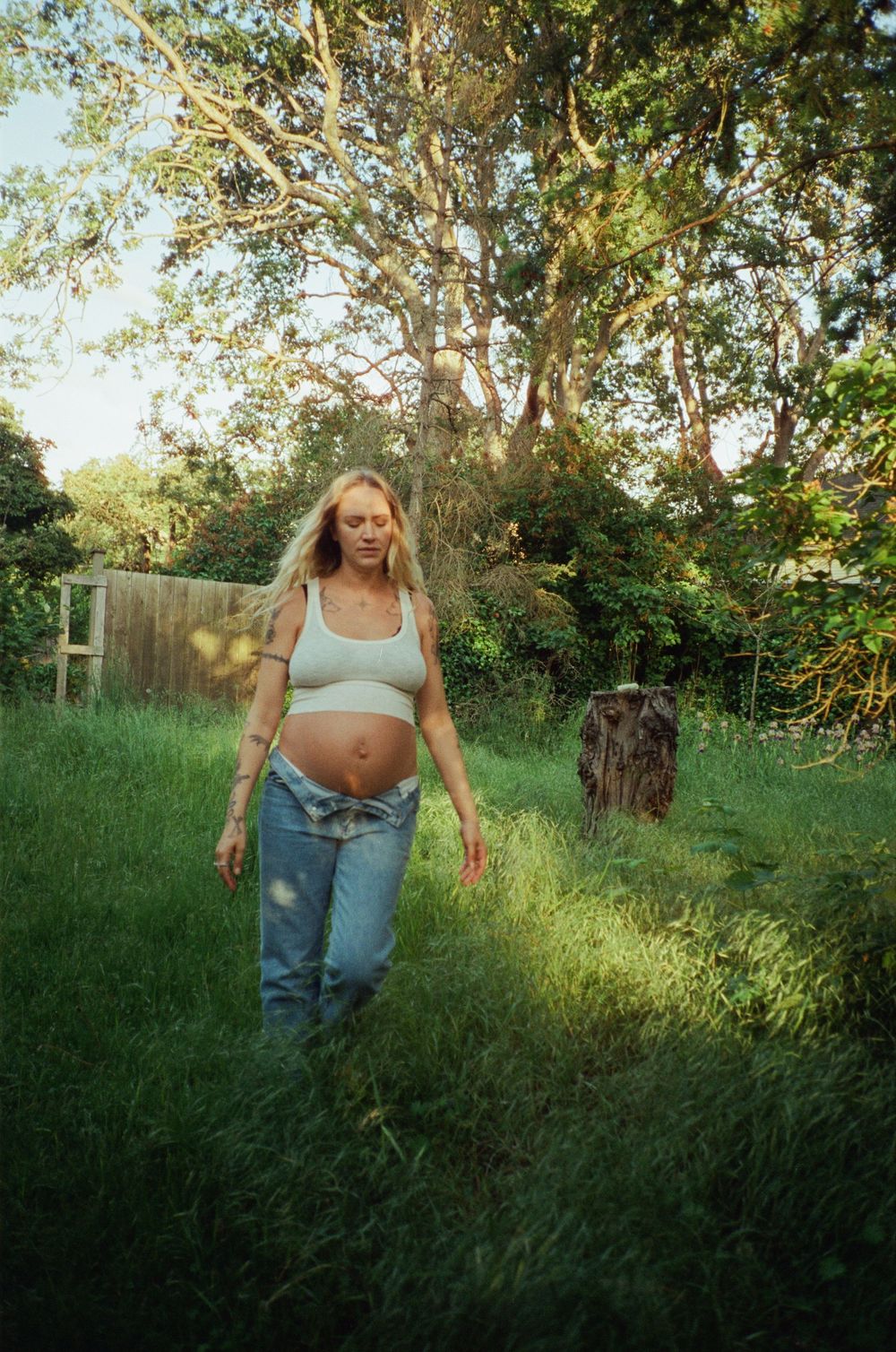 Pregnant blonde woman in jeans and crop top walking through a lush green garden at golden hour.