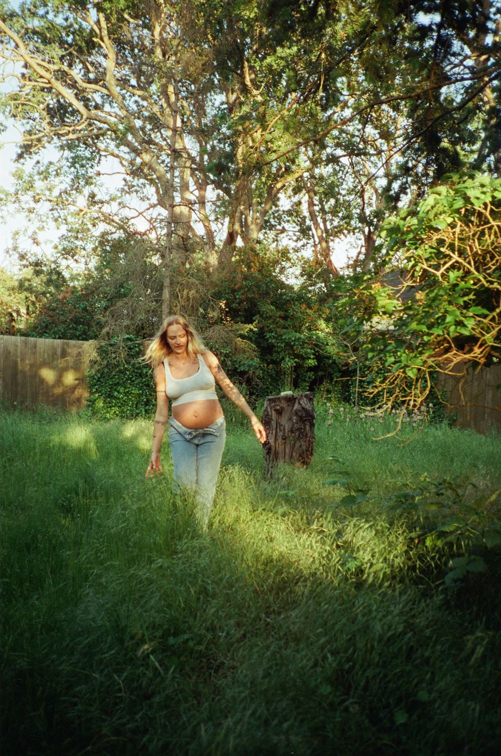 Pregnant woman walking through tall grass in a sunny backyard garden wearing jeans and crop top.