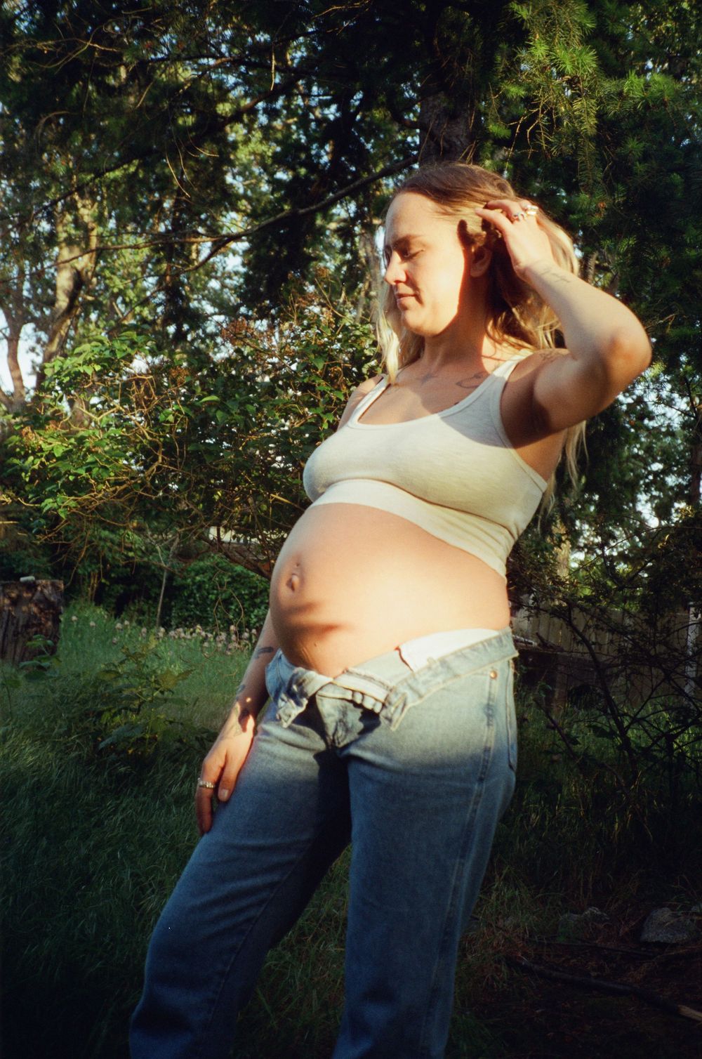 Pregnant woman in white crop top and jeans posing in a sunny garden with trees behind her.