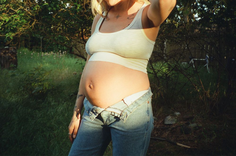 Close-up of a tattooed pregnant woman in a white crop top and low-rise jeans outdoors.