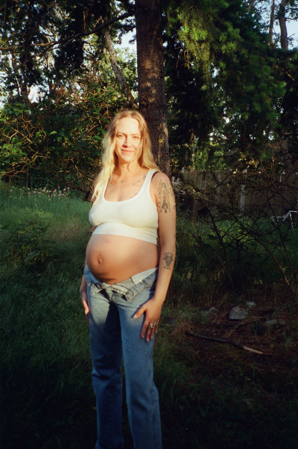 Smiling tattooed pregnant woman in white crop top and jeans standing in a green backyard.