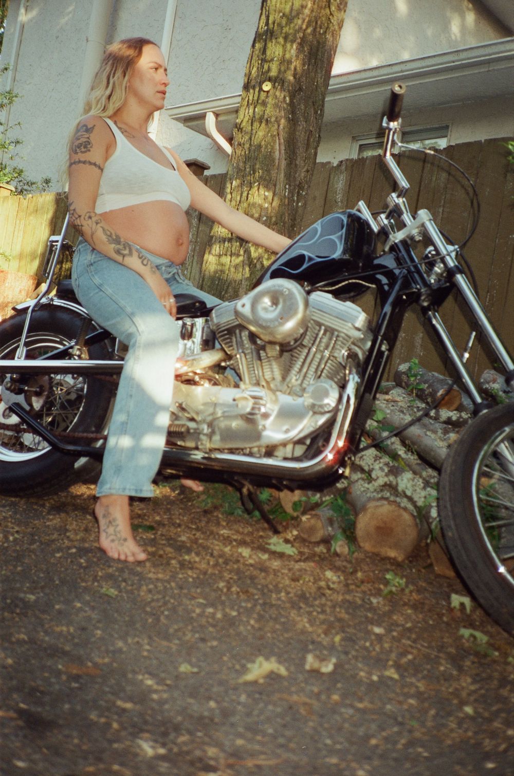 Tattooed barefoot pregnant woman posing beside a chrome chopper motorcycle in a backyard.