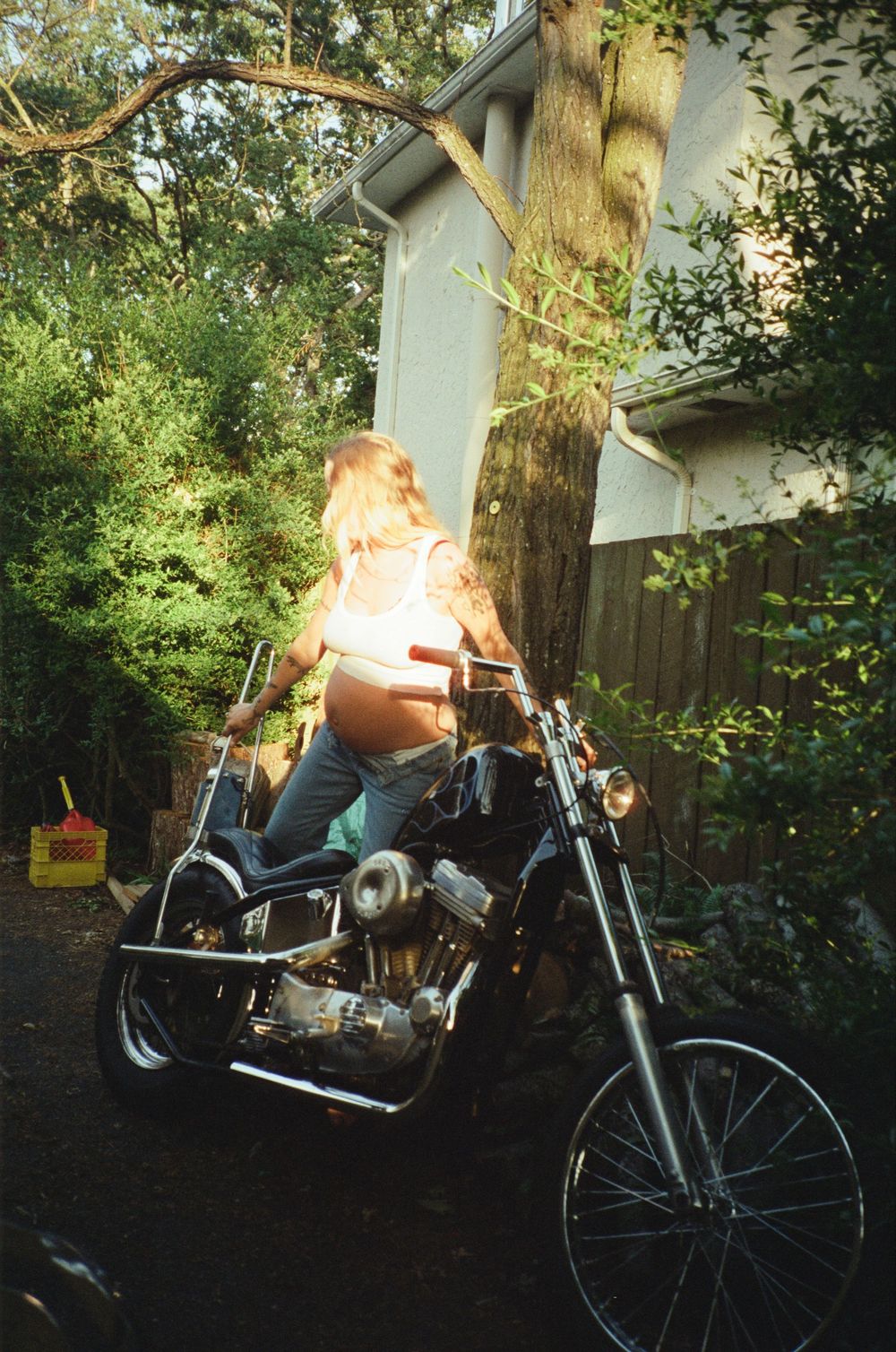 Pregnant woman sitting on a classic chopper motorcycle in a backyard surrounded by trees.