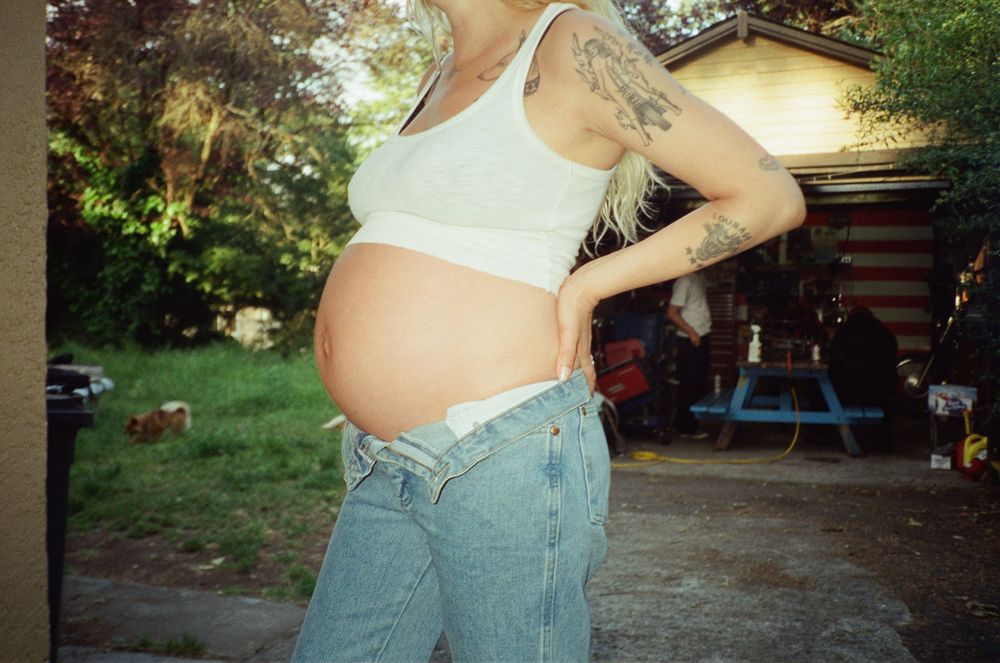 Tattooed pregnant woman in white crop top and low-rise jeans posing in a sunny backyard.