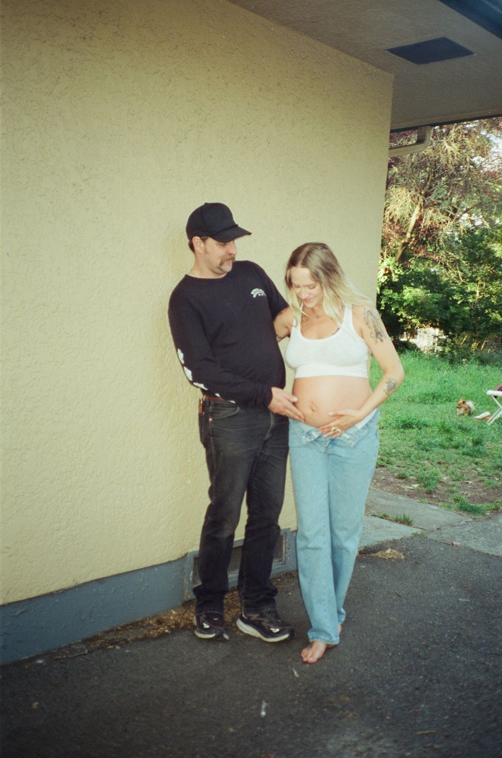Couple looking down at pregnant belly while standing against a yellow wall outdoors.