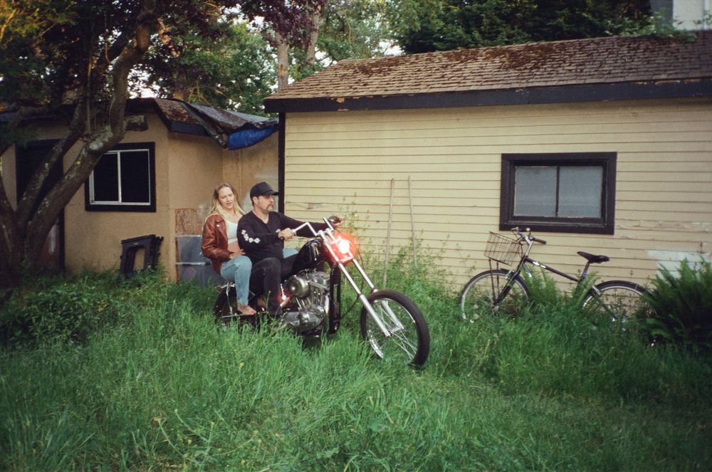 Two people sitting on a vintage motorcycle in an overgrown backyard near a yellow house.
