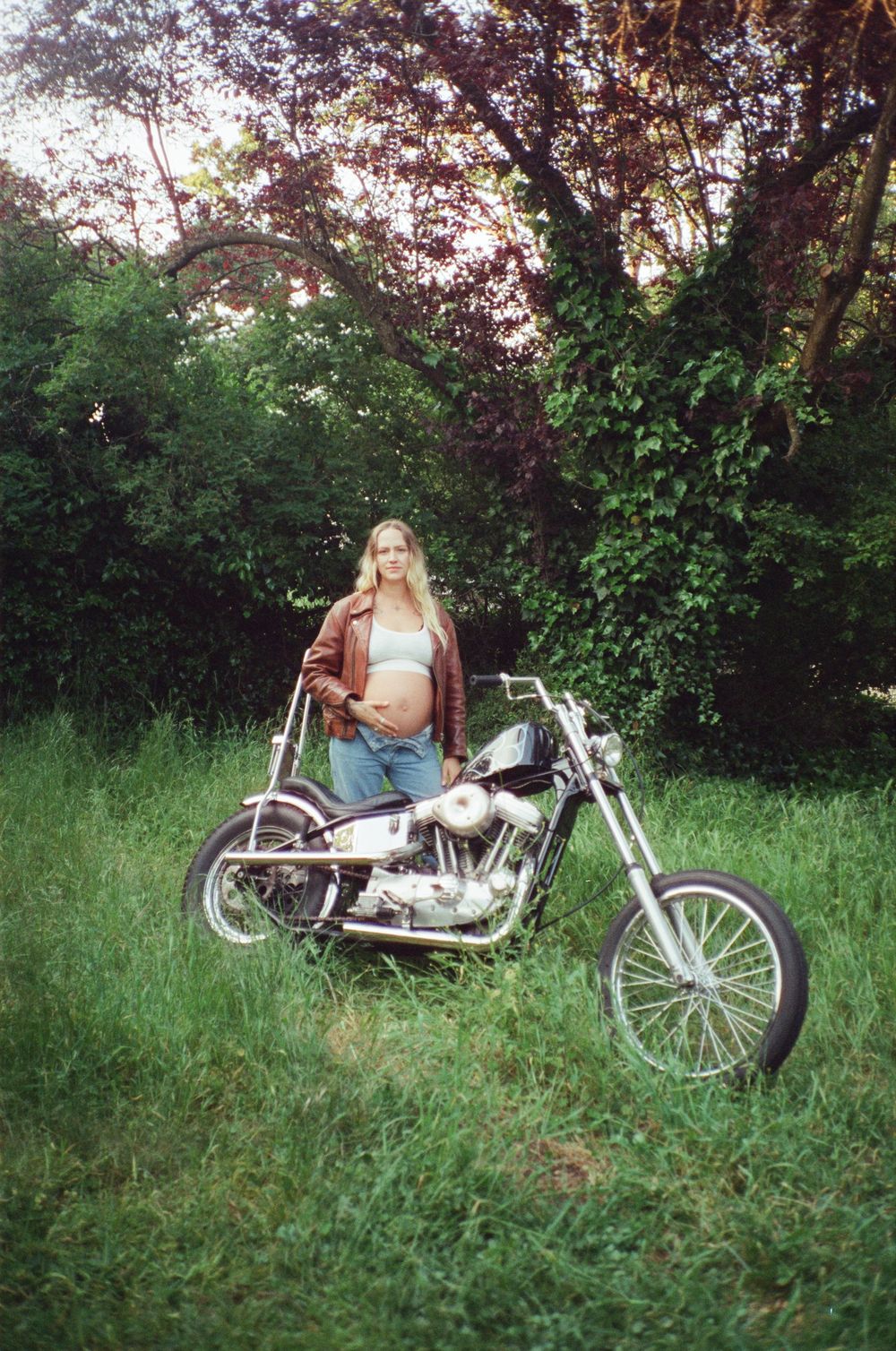 Pregnant woman in brown leather jacket standing beside a classic chopper in a lush green field.