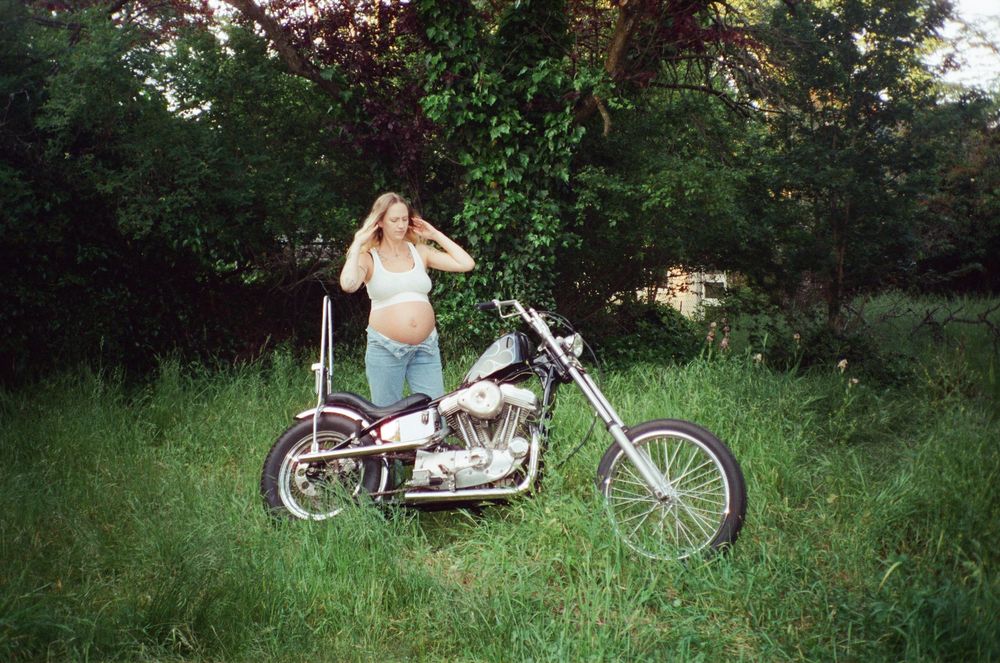 Pregnant woman standing behind a classic chopper motorcycle in a grassy outdoor setting.