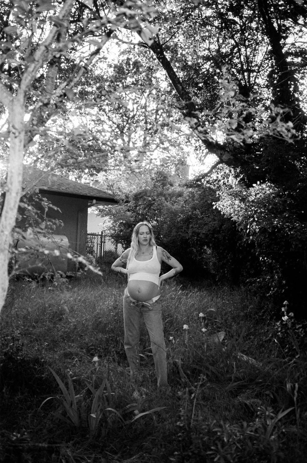 Black and white photo of a pregnant blonde woman standing in a lush backyard garden.