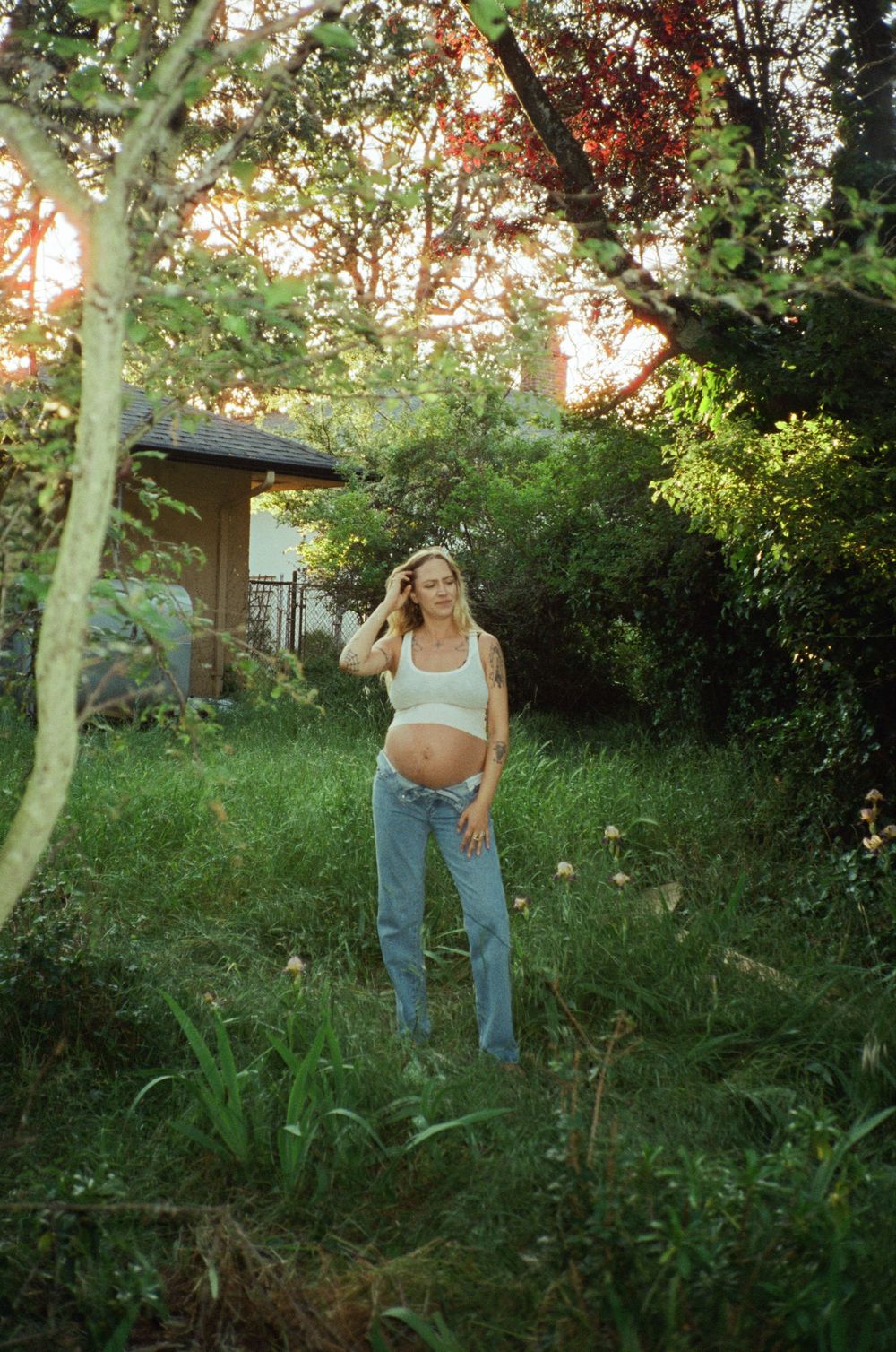 Pregnant blonde woman in jeans and crop top smiling in a sunlit green garden at sunset.