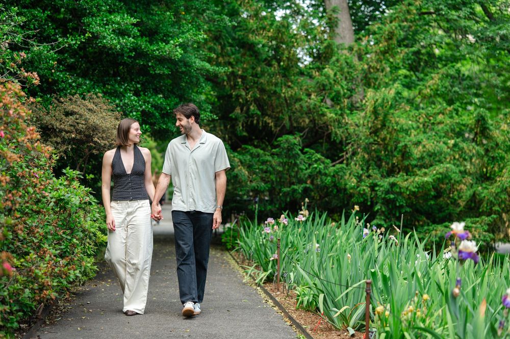 Couple holding hands walking along a garden path lined with green shrubs and iris flowers.