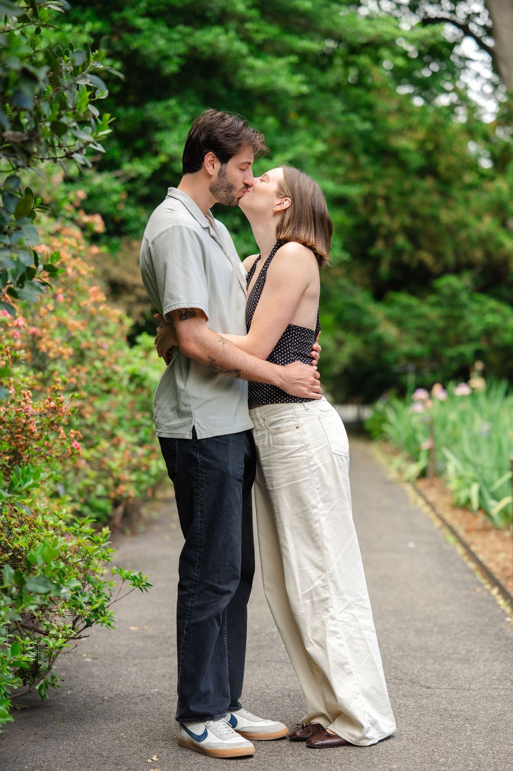 A couple sharing a romantic kiss on a garden path surrounded by lush green trees and colorful plants.