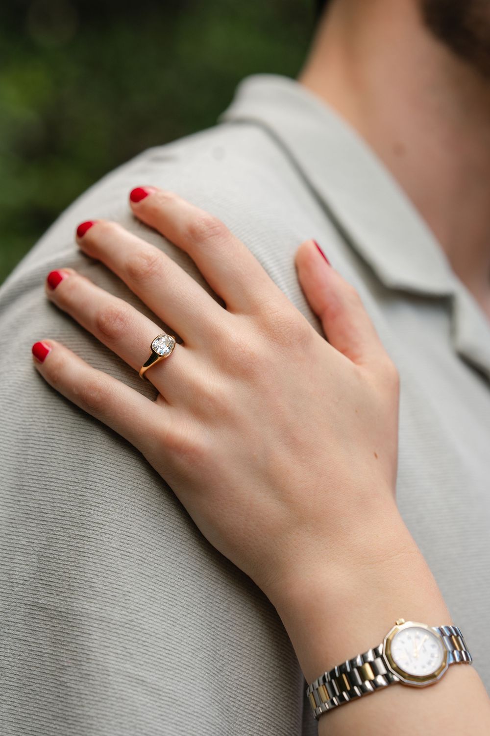 Woman's hand with red nails wearing a gold diamond solitaire ring and silver watch, resting on a shoulder outdoors.