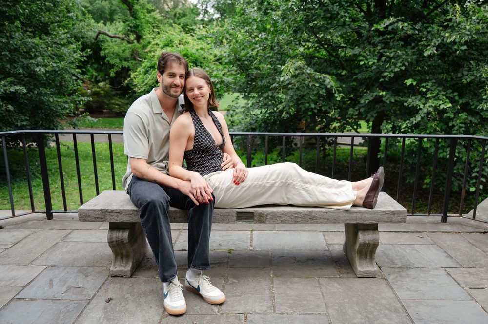 Couple sitting together on a stone bench outdoors, smiling, surrounded by lush green trees.