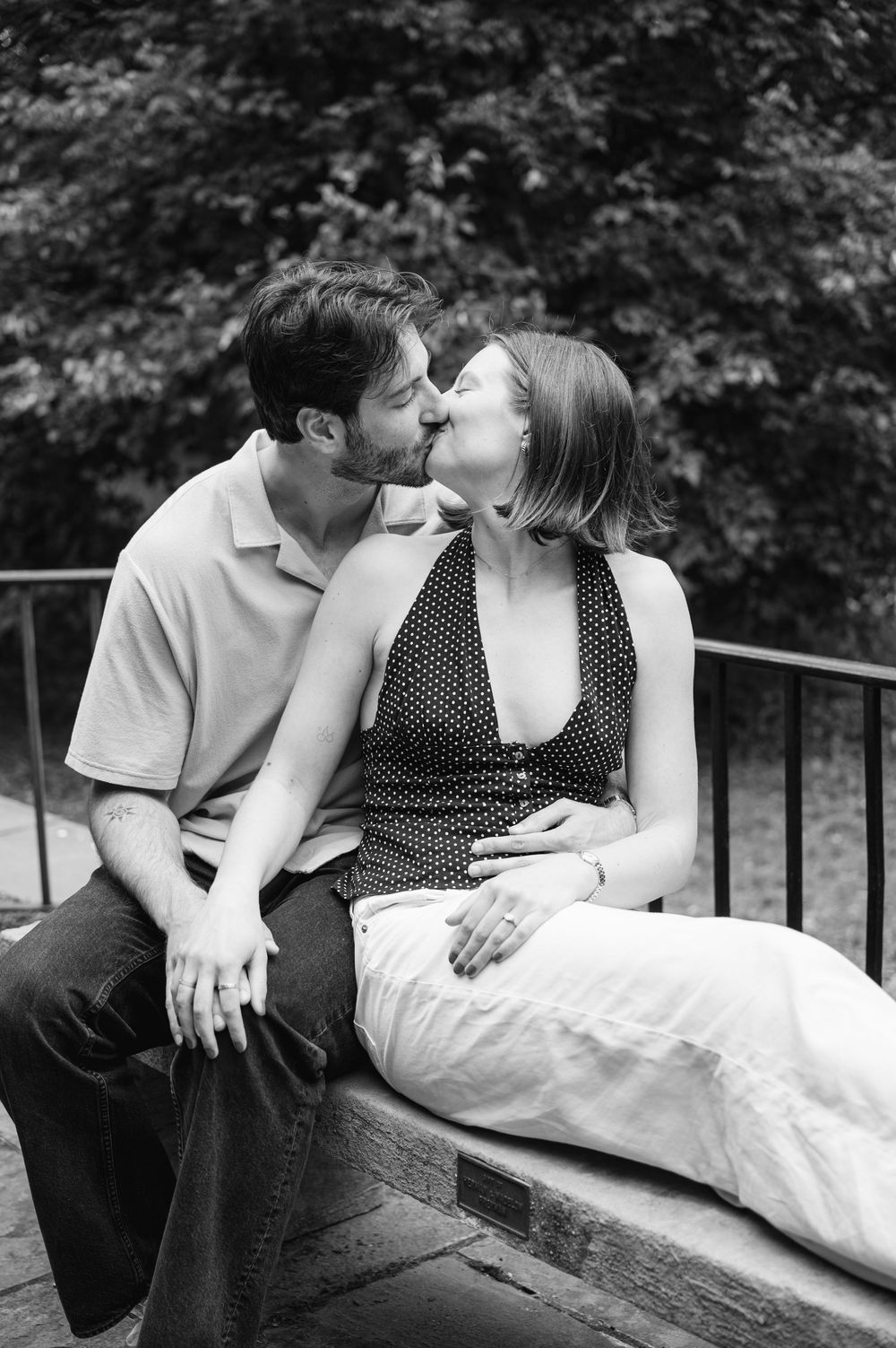 Black and white photo of a couple sharing a kiss while sitting on a bench in a lush park setting.