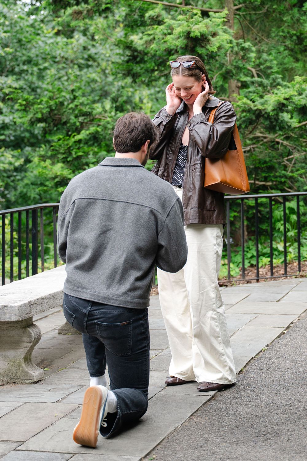 Man kneeling on one knee proposing to emotional woman in a park setting with lush green trees in background.