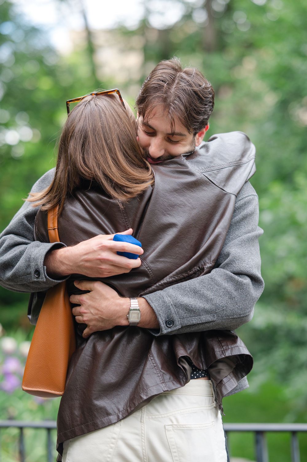 Couple embracing outdoors, woman holding blue ring box after marriage proposal, green trees in background.