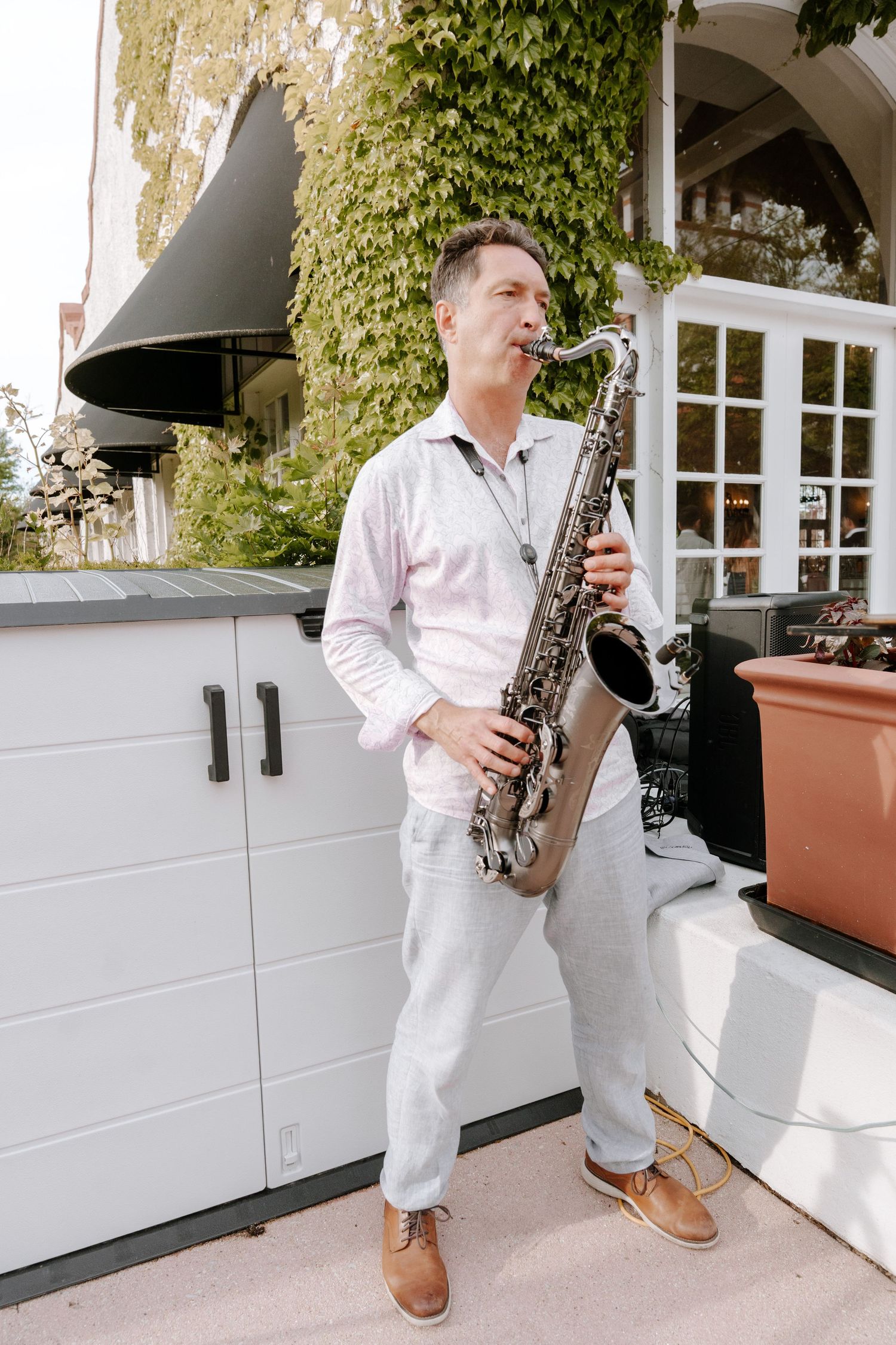 A man in white shirt and pants plays saxophone outdoors near ivy-covered building.