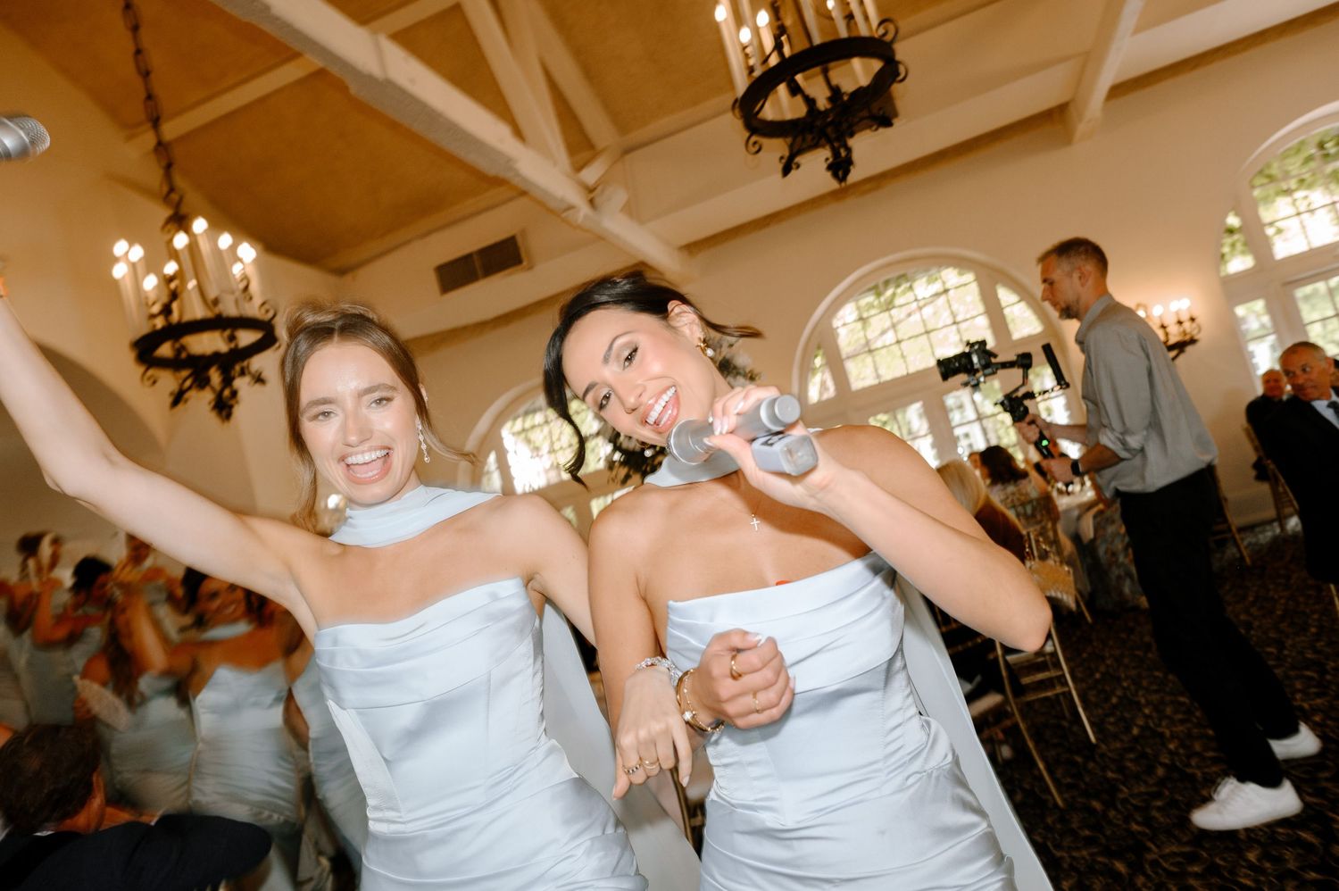 Two bridesmaids in white dresses dancing joyfully at an elegant wedding reception venue.