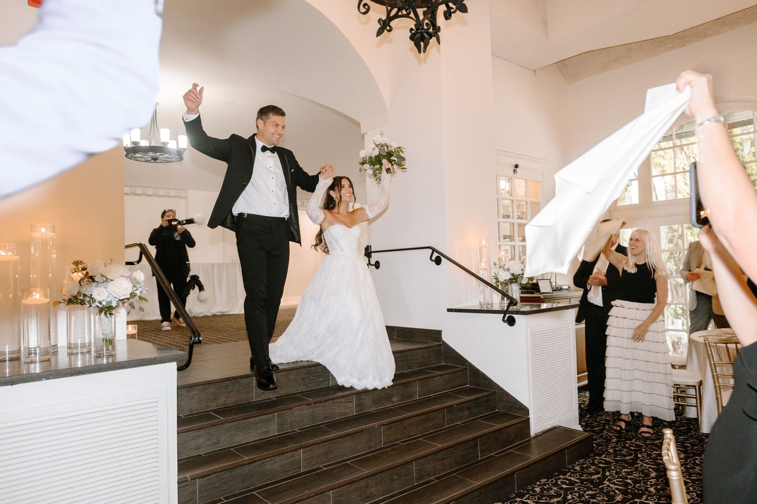 Bride and groom make grand entrance descending elegant staircase at wedding reception, guests celebrating around them.