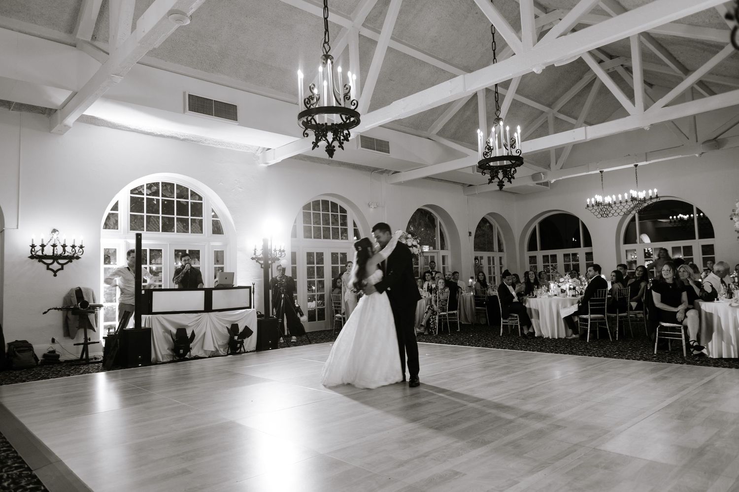 Bride and groom share first dance in elegant ballroom with chandeliers and seated wedding guests in background.