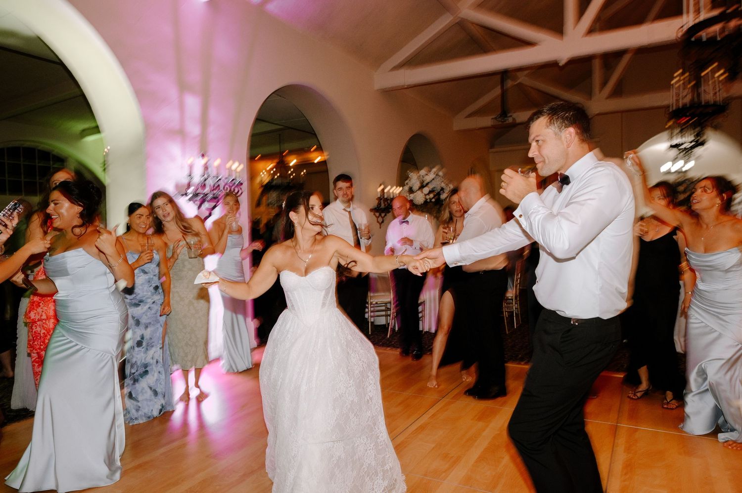 Bride and groom dancing joyfully with guests at elegant wedding reception on wooden dance floor.