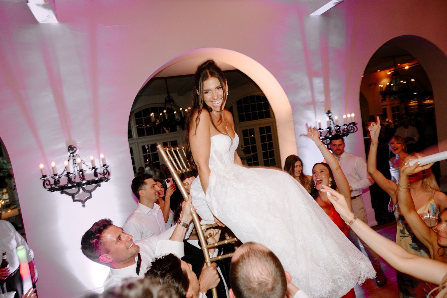 Bride in white gown lifted on chair during lively wedding reception celebration with guests cheering around her.