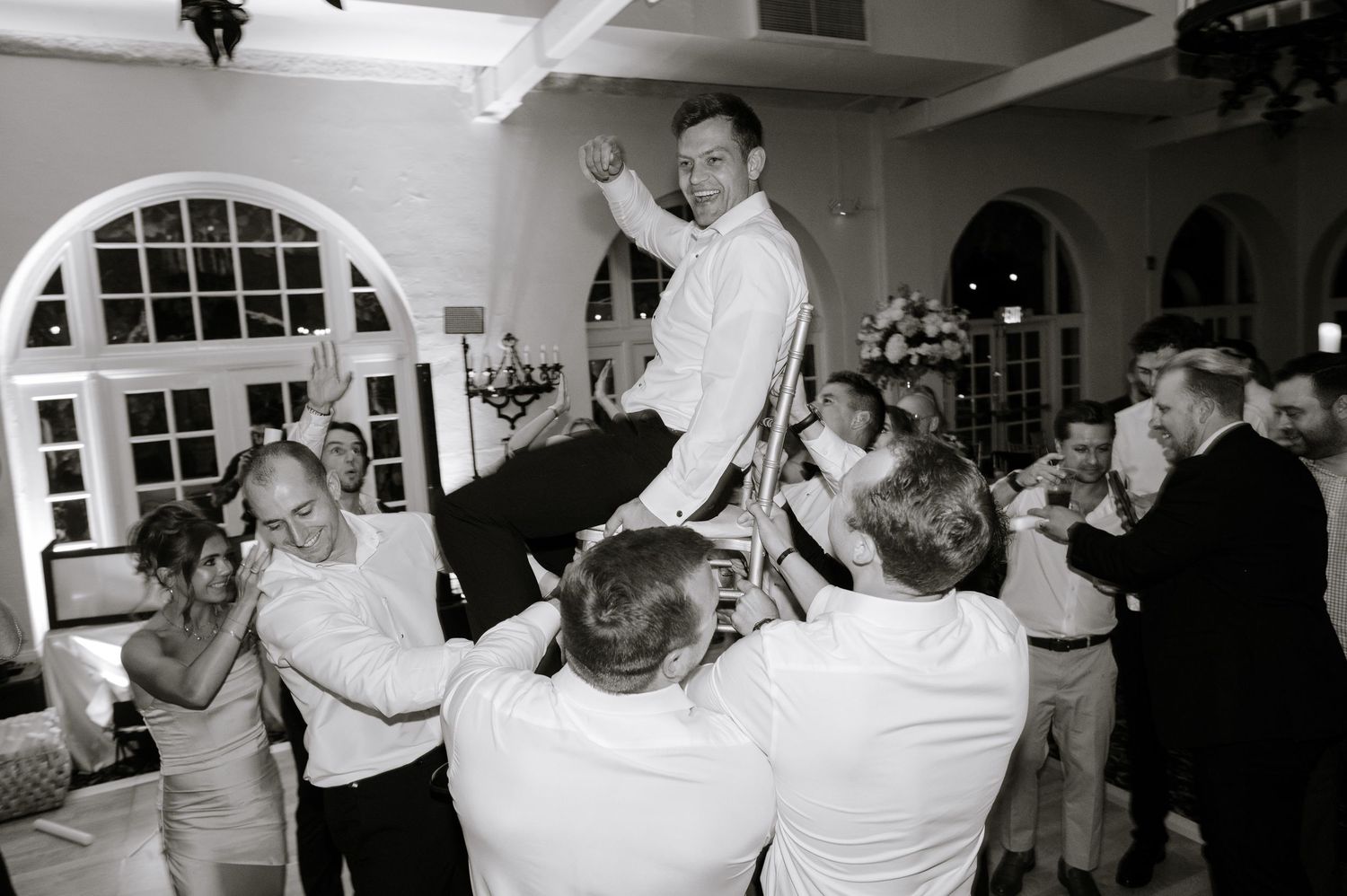 Groom lifted by groomsmen during wedding reception celebration in elegant white ballroom.