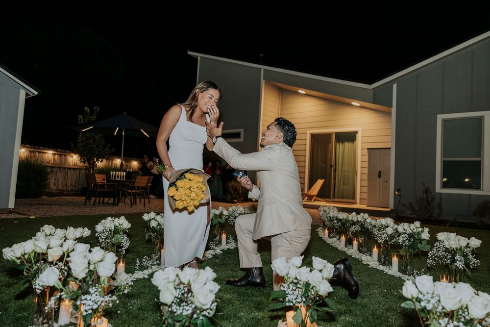 Man proposes to woman on one knee surrounded by white flowers and candles in a romantic backyard at night.