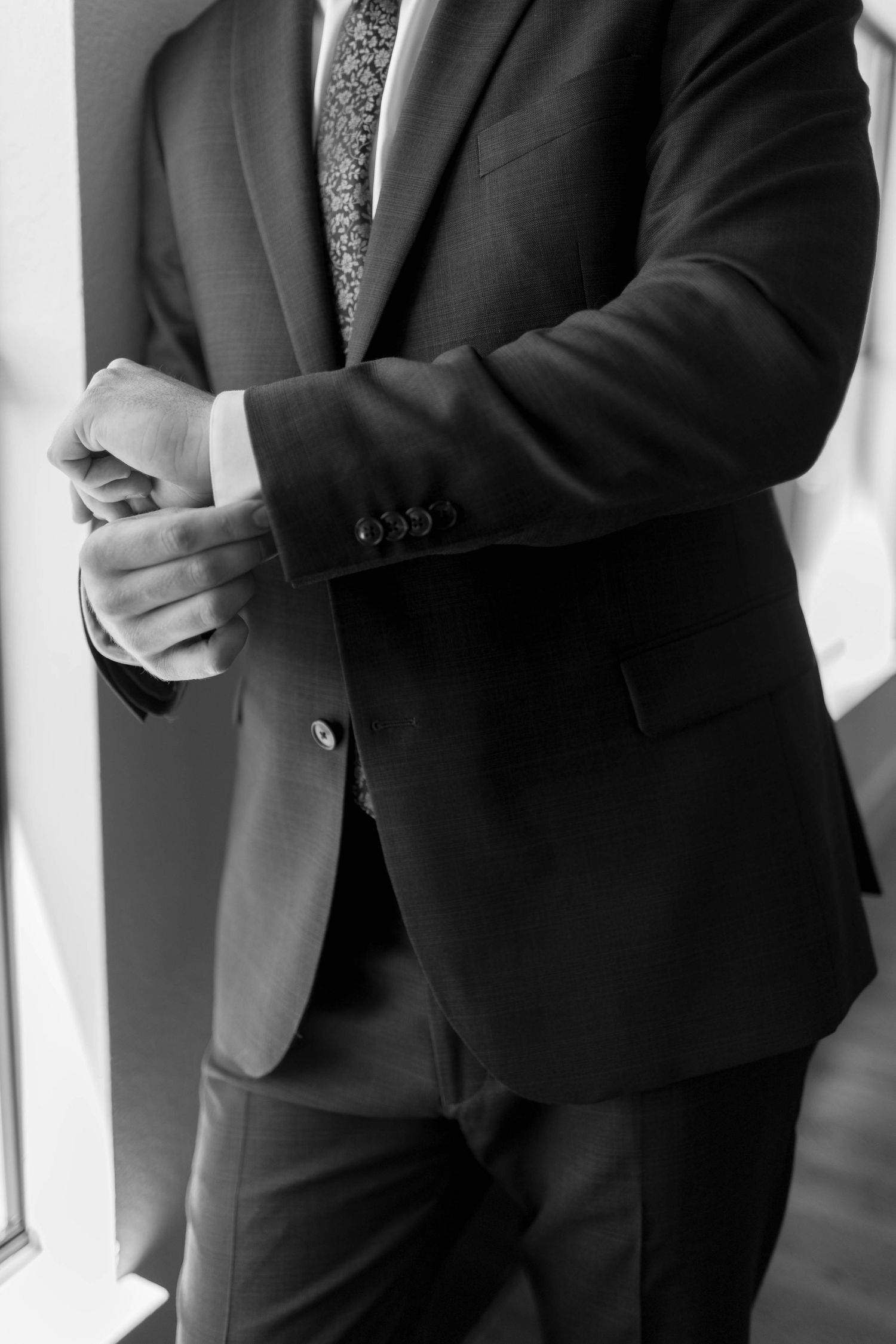 Black and white photo of a man in a dark suit adjusting his cufflinks, wearing a patterned tie.