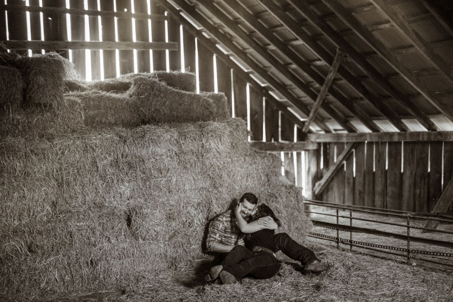 Black and white photo of a person resting against hay bales inside a rustic barn with wooden beams.