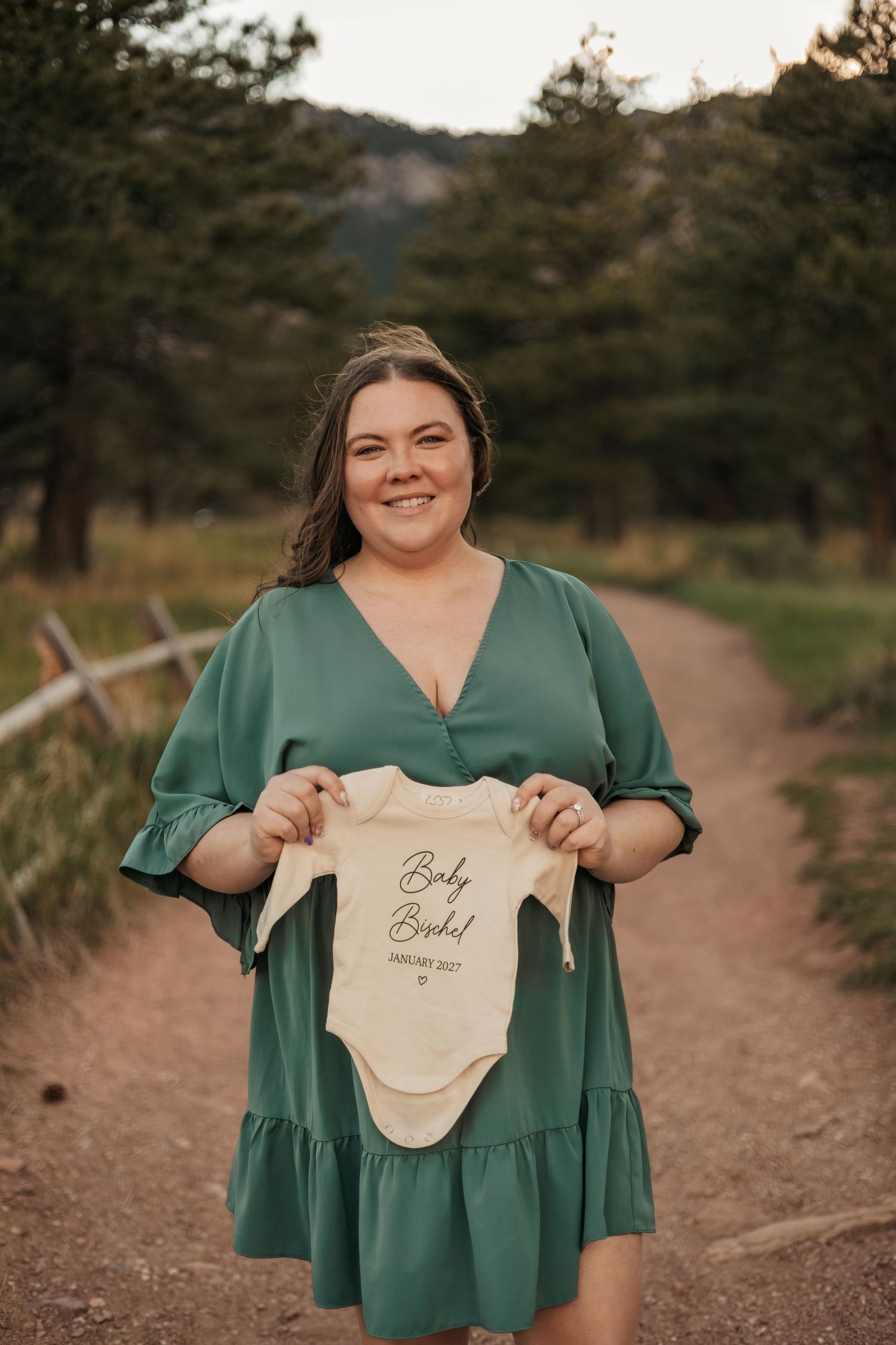 Smiling woman in green dress holding a baby onesie announcement | Baby Announcement Photographer in Boulder, Colorado