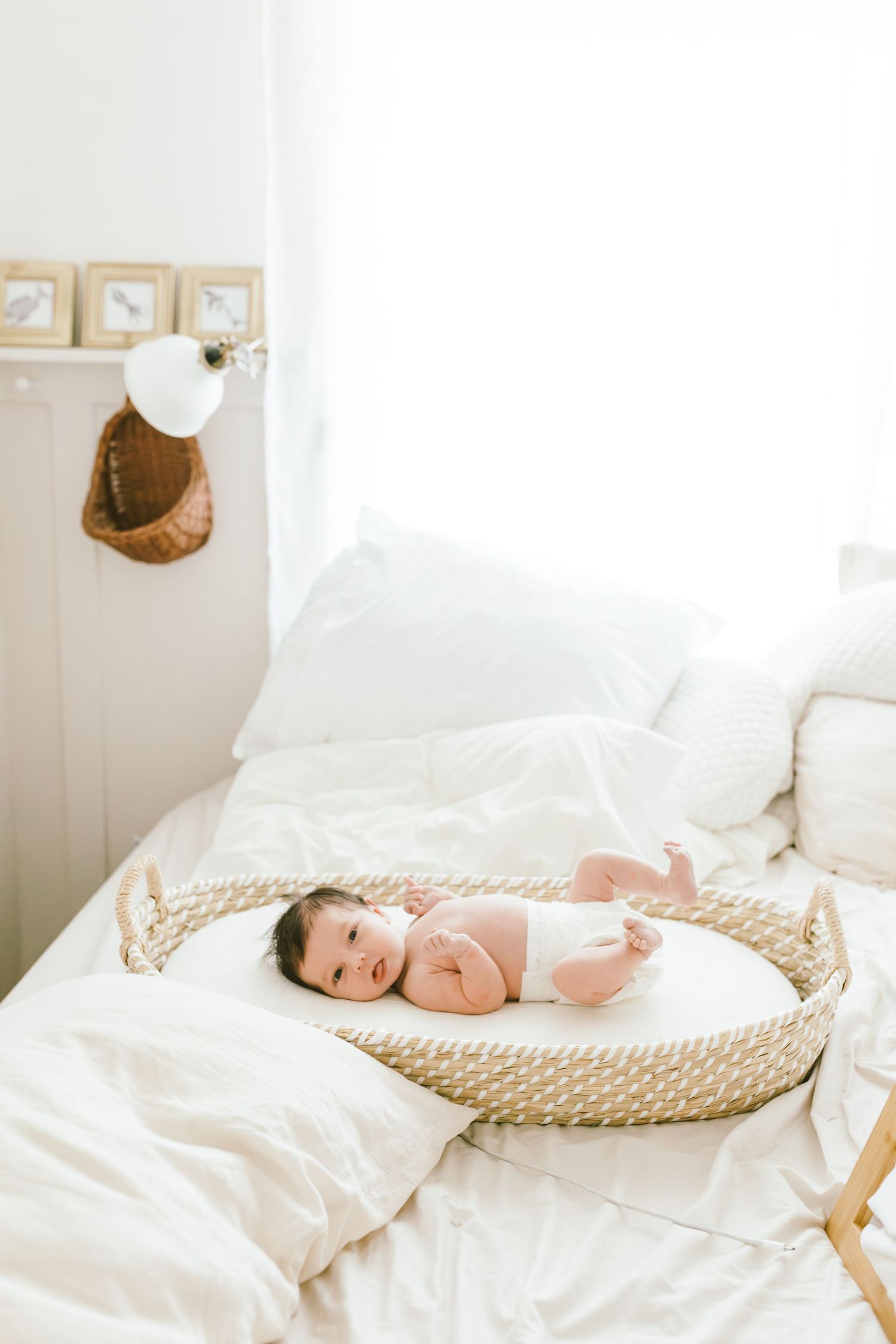 Newborn baby lying in a woven Moses basket on a white bed in a bright, airy nursery room.
