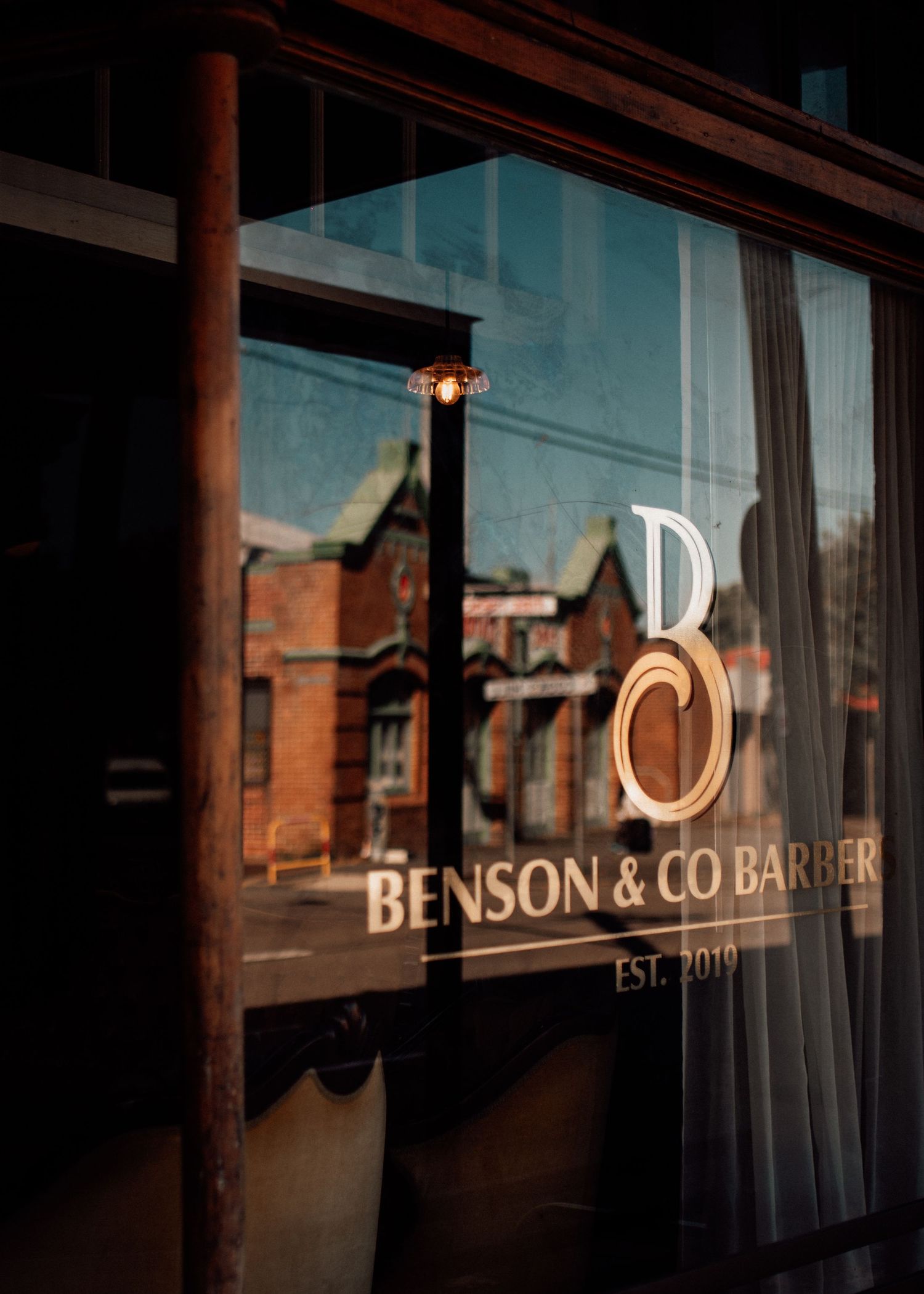 Benson & Co Barber shop window sign established 2019, reflecting a brick building street view in warm golden light.
