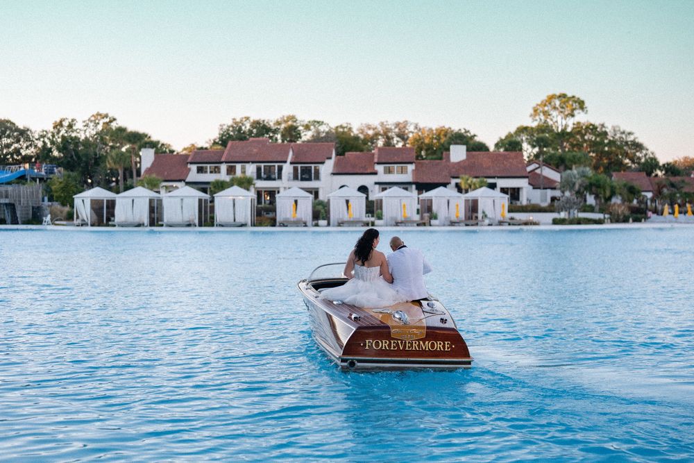 Couple riding a wooden boat on a blue lagoon with white Mediterranean-style villas in the background.