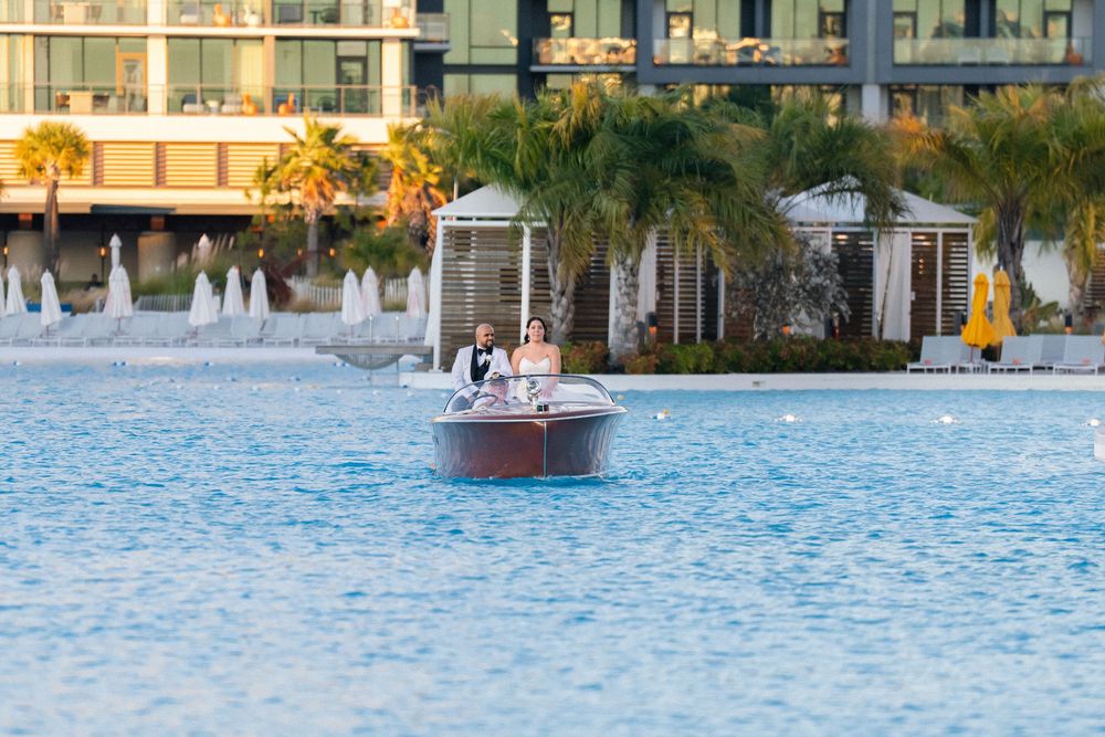 Couple in wedding attire riding a wooden boat on a blue lagoon with palm trees and resort buildings behind them.