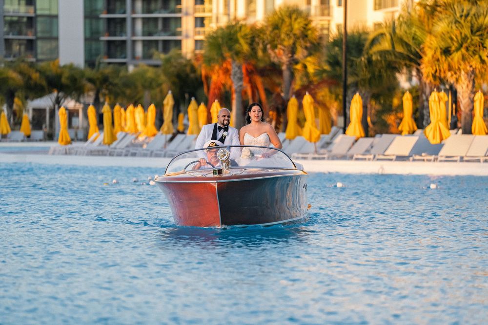 Bride and groom riding a classic wooden boat on a resort pool with yellow umbrellas and palm trees in the background.