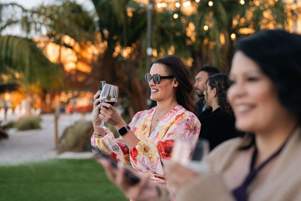 Woman in floral dress smiling while holding a camera outdoors at golden hour with palm trees in background.