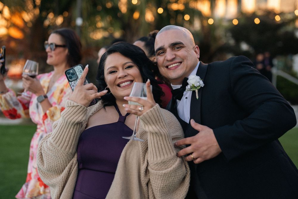 Couple smiles and poses with wine glasses at a festive outdoor wedding celebration at sunset.