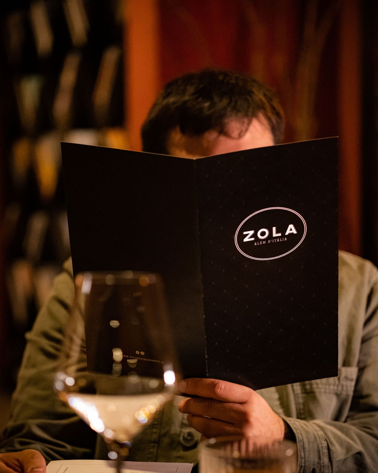 A diner reads a black Zola Wine & Spirits menu at a dimly lit restaurant table with a glass of white wine nearby.