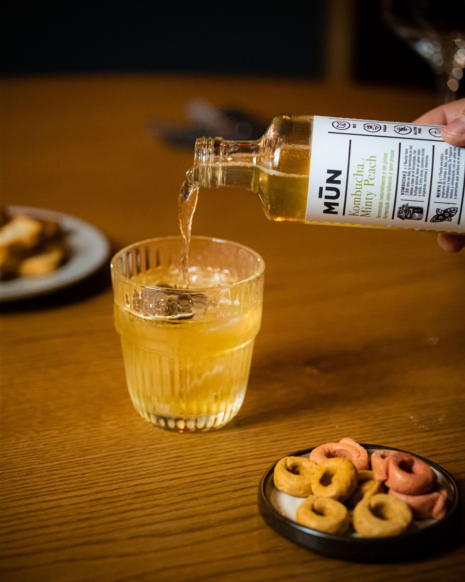 A hand pouring MUN kombucha into a ribbed glass on a wooden table with colorful ring snacks nearby.