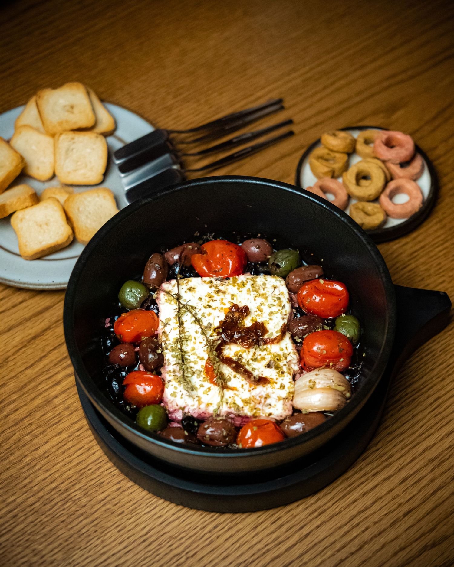 Black pan with baked feta, cherry tomatoes, olives, and herbs served with bread cubes and ring crackers.