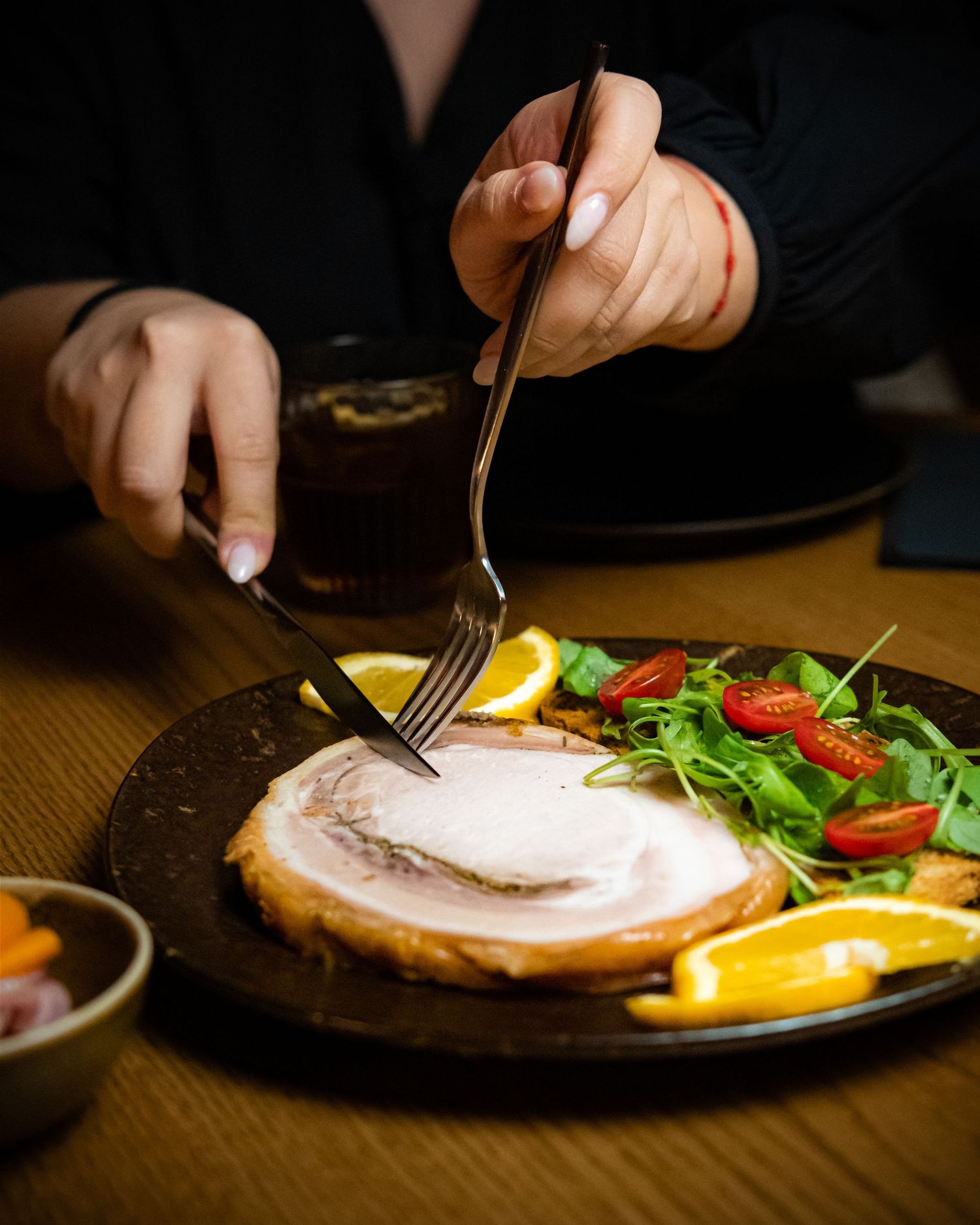 Woman cutting a juicy ham steak with fork and knife on a dark plate with fresh salad, tomatoes, and orange slices.