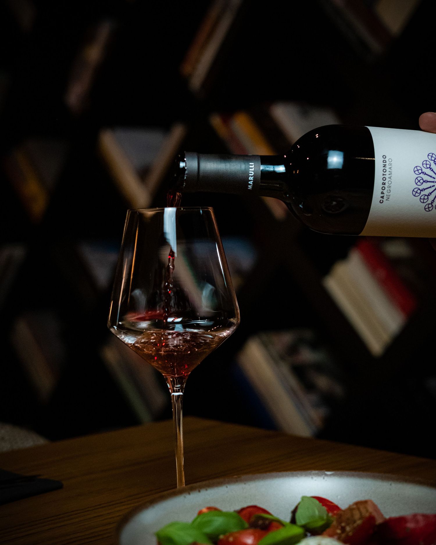Red wine being poured into an elegant crystal glass beside a fresh salad plate in a dimly lit wine cellar.